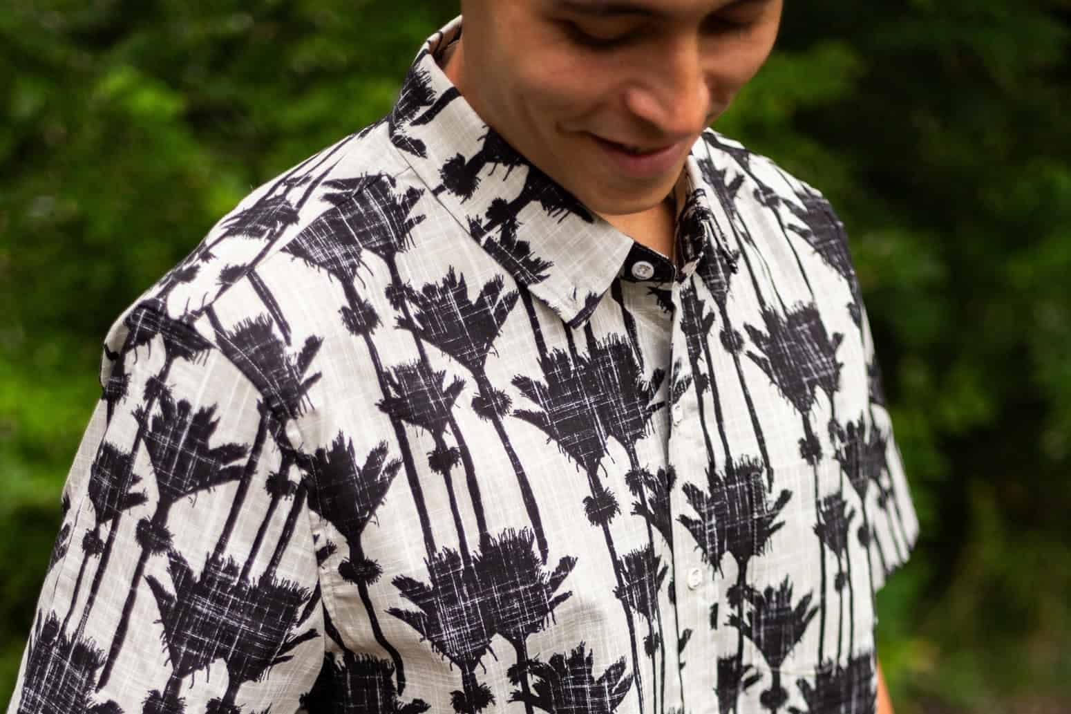 Kaleo-art-to-wear-17 | Must Do New Zealand | Must Do New Zealand Man wearing a white shirt with a black palm tree pattern, outdoors with greenery in the background