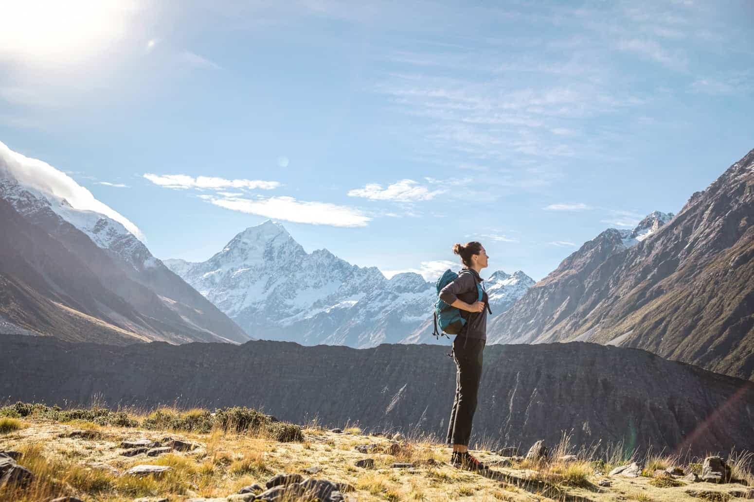 Cheeky-kiwi-tasman-Glacier-mount-Cook-9 | Must Do New Zealand | Must Do New Zealand Hiker with a teal backpack stands on a grassy rocky foreground, with snow?capped mountains and a clear blue sky behind.