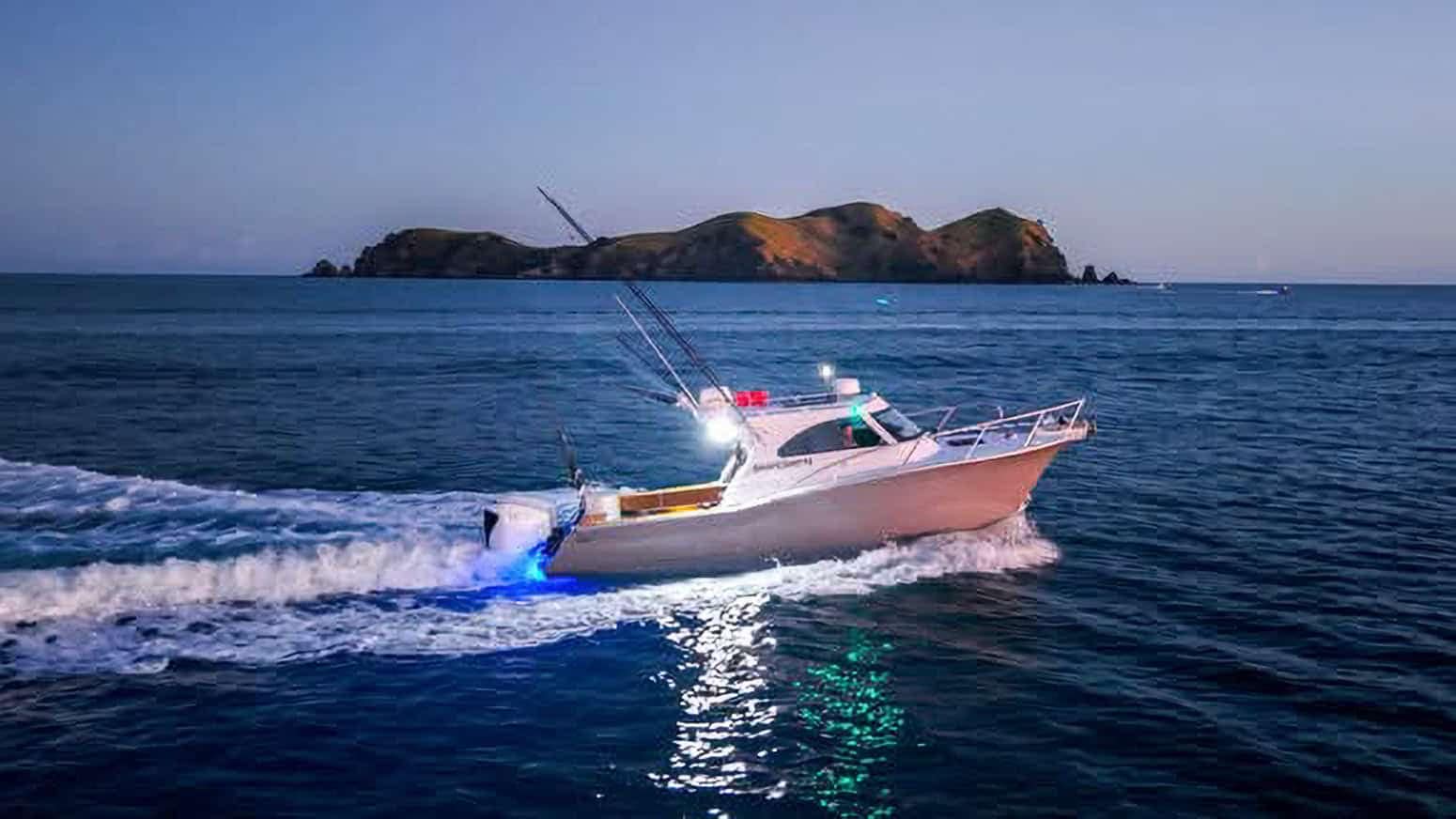 Days-Out-Fishing-Bay-of-Islands-15 | Must Do New Zealand | Must Do New Zealand White motorboat speeding across blue ocean, island in distance, with wake and deck lights reflecting on the water.