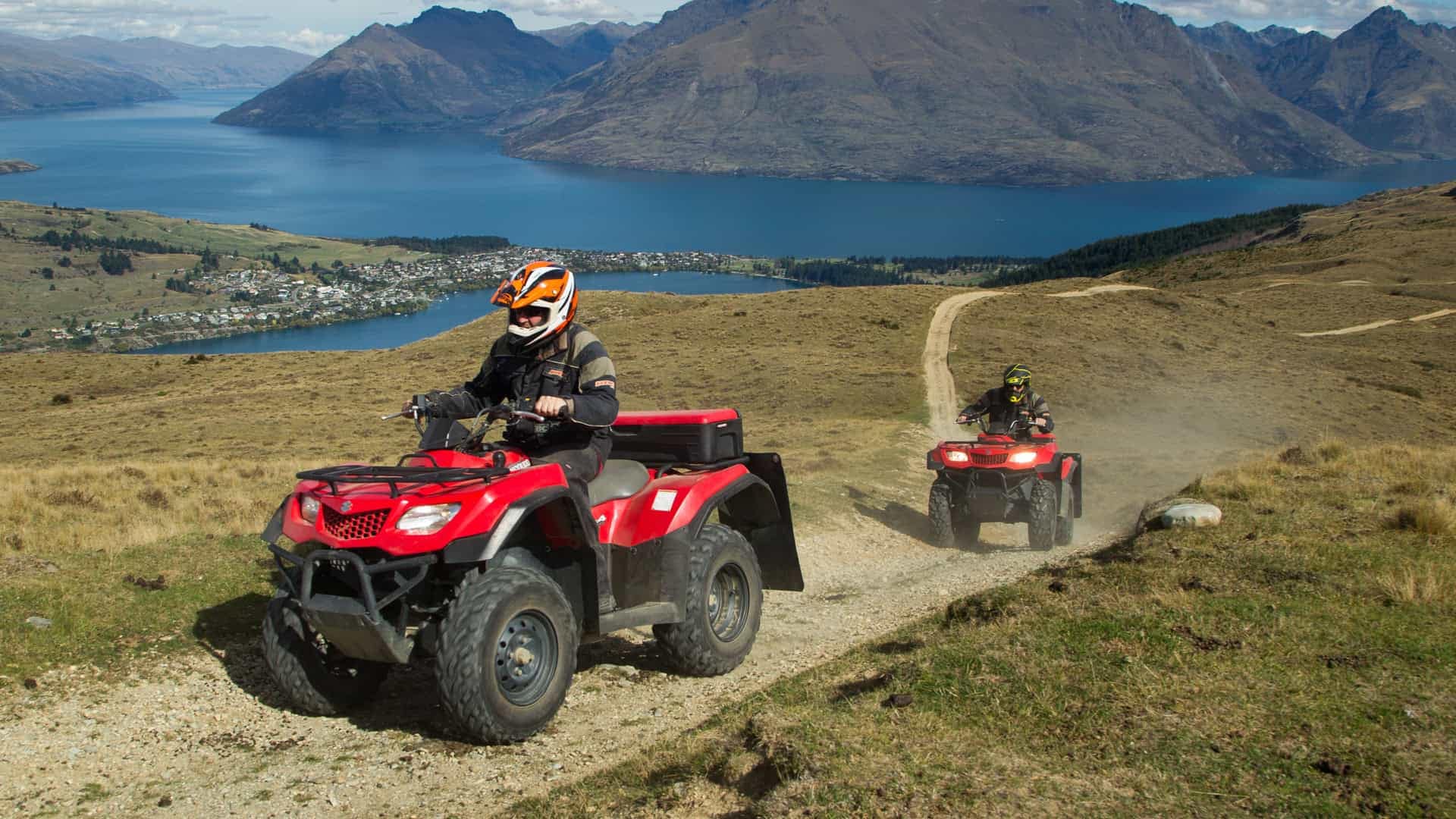 Nomad-safaris-quad-bike-tours-queenstown-1 | Must Do New Zealand | Must Do New Zealand Two riders on red ATVs travel a dusty hillside trail with a blue lake and towering mountains in the background.